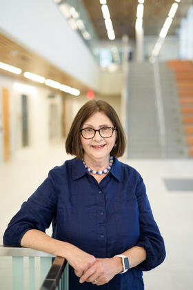 A portrait of Barb Smith in a dark blue blouse posing with a modern landing and staircase behind her