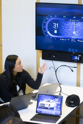 Students gather in an event space around TVs and tablets displaying designs for various automotive UX interface applications.