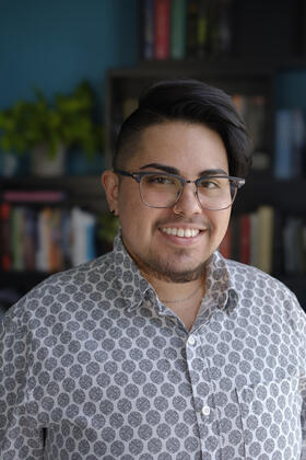 A smiling latiné person with dark swept-back hair, wire glasses, and a short beard, wears a geometric patterned button up shirt, while standing in front of a colorful bookshelf. 