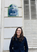 Amelia Acker, a person with shoulder length dark hair and glasses, wearing a dark blue jacket, smiling. Amelia is standing in front of a building with a blue and green sign reading "Rue des Archives."