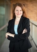 A woman with red hair, wearing a black suit and blue shirt, smiling. 