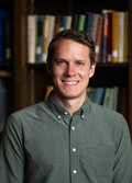 Kevin Carney, a person wearing a dark blue/green button up shirt, with dark hair, smiling. There is an out-of-focus bookshelf with books in the background.  