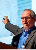David Lazer, pointing to an image on a projection screen, wearing a suit jacket, blue shirt and glasses. 