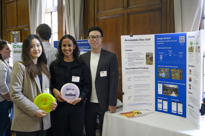 Three students standing next to a tri-fold poster titled "Accessible Disc Golf." Two of them are holding discs