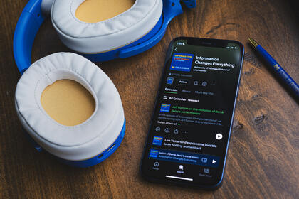 Blue and yellow headphones rest on a dark wood background beside a phone with the Information Changes Everything podcast displayed on Spotify. The latest episode is highlighted.