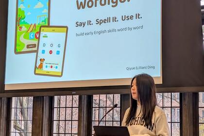 A student presents at a podium in front of a slide that reads "Wordigo: Say it. Spell it. Use it."