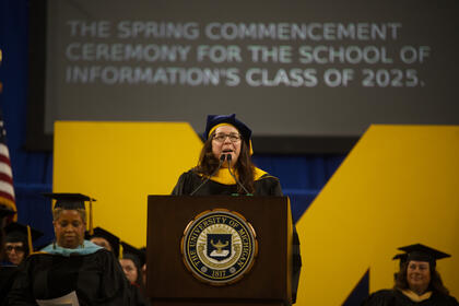 Andrea Forte speaks at a podium on a stage with the Block M visible behind her. A transcript on the screen reads "The spring commencement ceremony for the School of Information's Class of 2025."