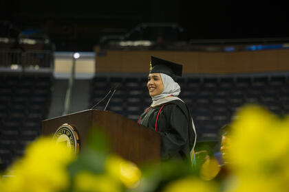A close-up side view of Hibah Al-Jabur speaking at a podium, with out-of-focus yellow flowers in the foreground