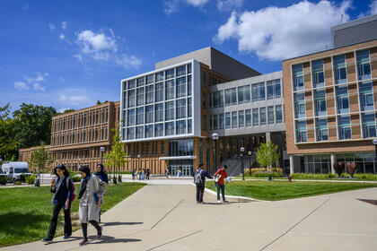 A far view of the Leinweber Building with scattered students walking the sidewalk in front of them, and two of them greeting each other by bumping fists