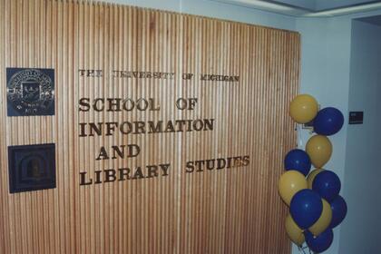 A wall sign reads School of Information and Library Studies with maize and blue balloons nearby.