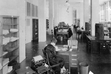 A black and white photo of a large room with desks, typewriters and bookshelves. There are a few people at the desks.
