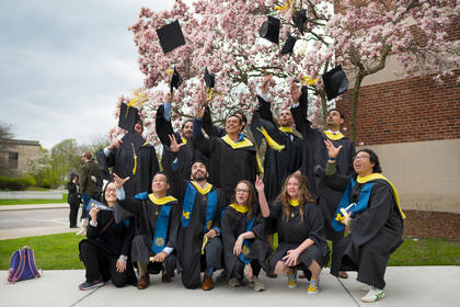 A group of graduates in gowns, tossing their caps in front of a pink flowering tree