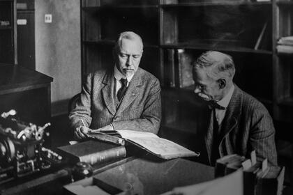 William Warner Bishop sits at a table with a colleague looking over a book near a wall of barrister bookcases.