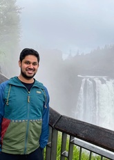 Krishna Vadrevu stands on a bridge in front of a roaring waterfall. A forest of pine trees on the horizon is partially obscured by fog.