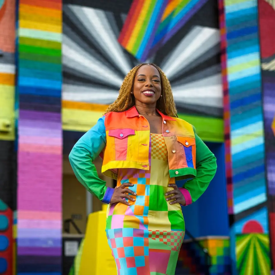 Megan poses in front of a rainbow and prismatic mural in an alleyway while wearing a vibrant dress and jacket of a patchwork of shapes and colors.