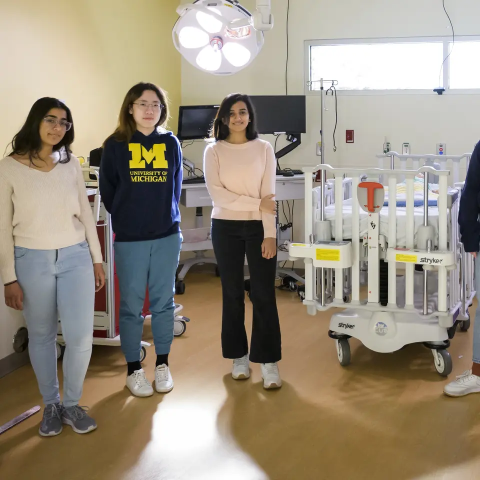 A photo of five students standing in a light-filled operating room. Two of them wear "University of Michigan" sweatshirts.